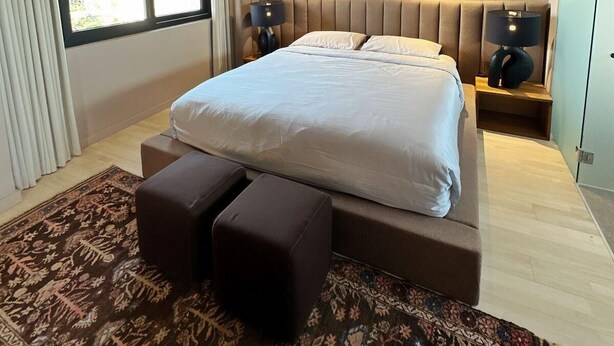 Un dormitorio moderno con una cama grande, cabecera, dos mesitas de noche con lámparas, una alfombra estampada y vistas a través de una ventana.