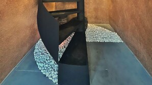 Una escalera de caracol con barandilla negra y peldaños, rodeada de paredes con textura y un suelo con diseño de guijarros.