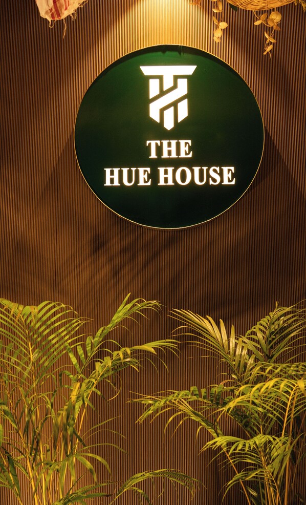 A sign with the text "THE HUE HOUSE" on a green background, surrounded by vertical wooden slats and some greenery at the bottom. - The Hue House (Sivasagar)