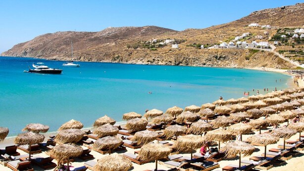 A beach with rows of sun loungers and thatched umbrellas, a clear blue sea, and buildings on a hillside in the background. - Eidolion Essence Nest - Mykonos Cozy Getaway (Mykonos)