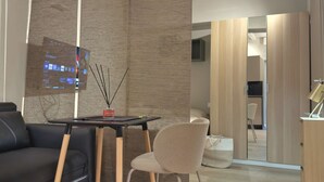 Una sala de estar moderna con un sofá negro, un sillón color beige, una mesa de centro de madera y un televisor montado en una pared con textura.