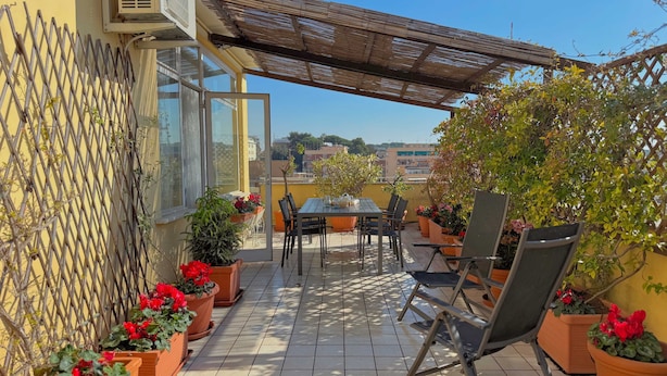 Un patio con mesa y sillas, rodeado de plantas en macetas y una pérgola de madera.