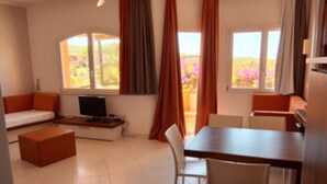Ein Zimmer mit einer Couch, einem Fernseher, einem Esstisch mit Stühlen und großen Fenstern mit Vorhängen.