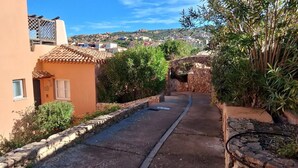 Eine enge Gasse mit Steinwänden und viel Grün, die zu einer Anhöhe mit Gebäuden führt.