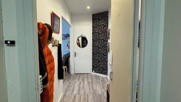 Een gang met een houten vloer, een kapstok met een jas, een deur met een spiegel en een wand met een bladmotief.
