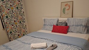 A neatly made bed with striped bedding, a rolled towel, and slippers on the bed. There are pillows with striped and patterned designs. A colorful curtain with a floral pattern is partially visible on the left. - Large, bright bedroom (Molenbeek)