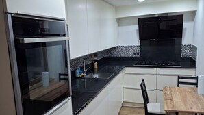 A modern kitchen with white cabinets, a black countertop, a built-in oven, and a black range hood. There is a dining area with a wooden table and chairs. - Large, bright bedroom (Molenbeek)