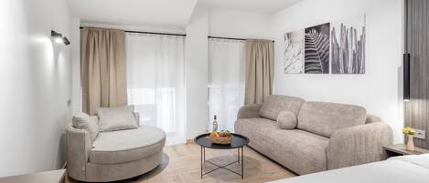 A modern, well-lit living room with a sofa, a round chair, a small table with a fruit basket, and artwork on the walls.