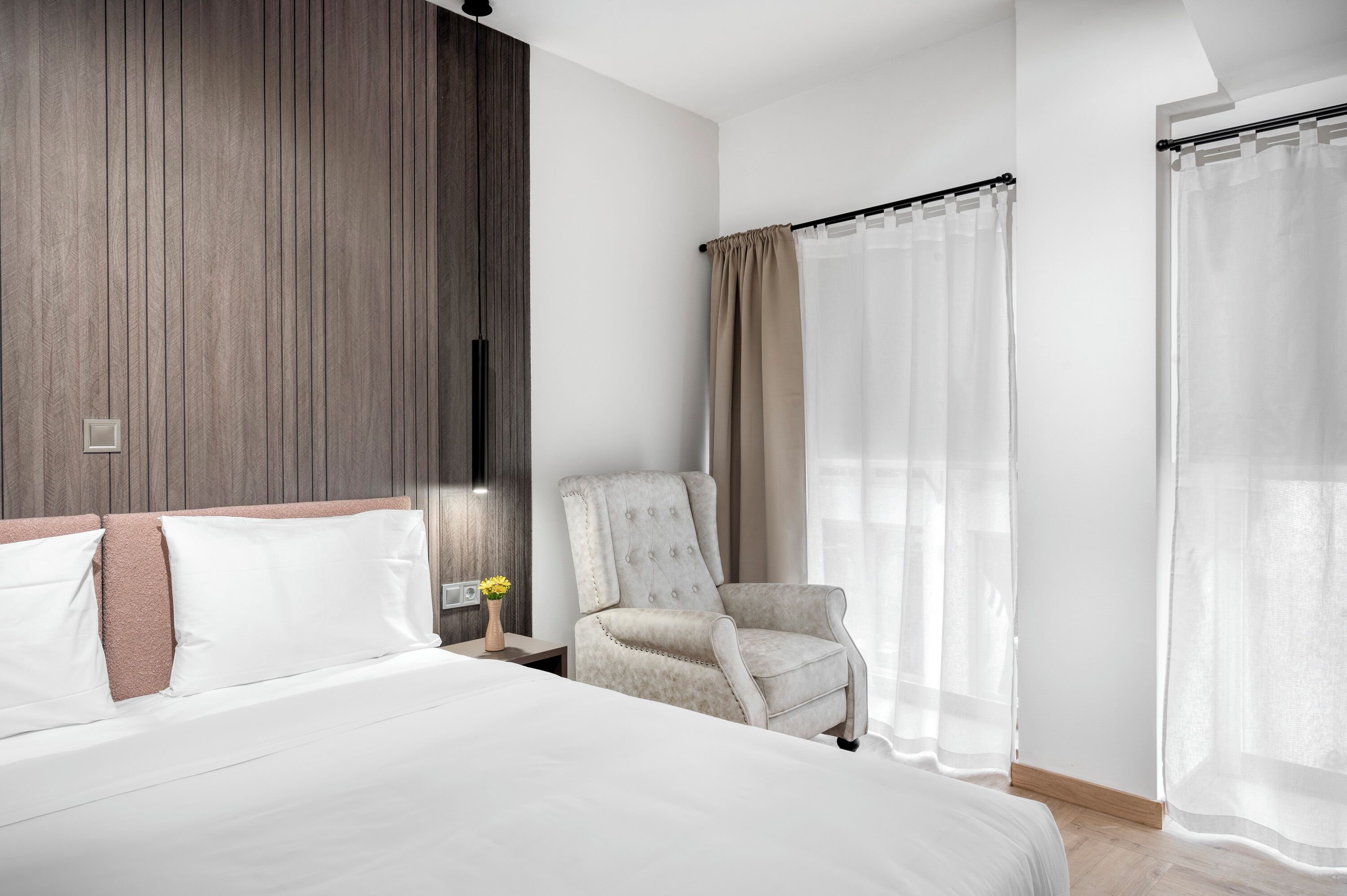 A hotel room with a large bed, a chair, a nightstand with a vase, and sheer curtains.