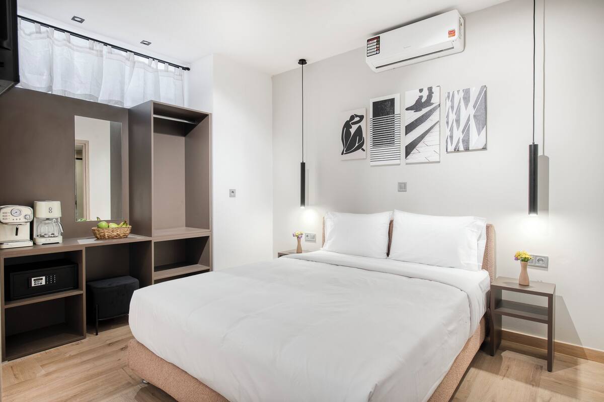 A modern hotel room with a large bed, a wardrobe, a mirror, artwork on the wall, and a bedside table with a lamp.