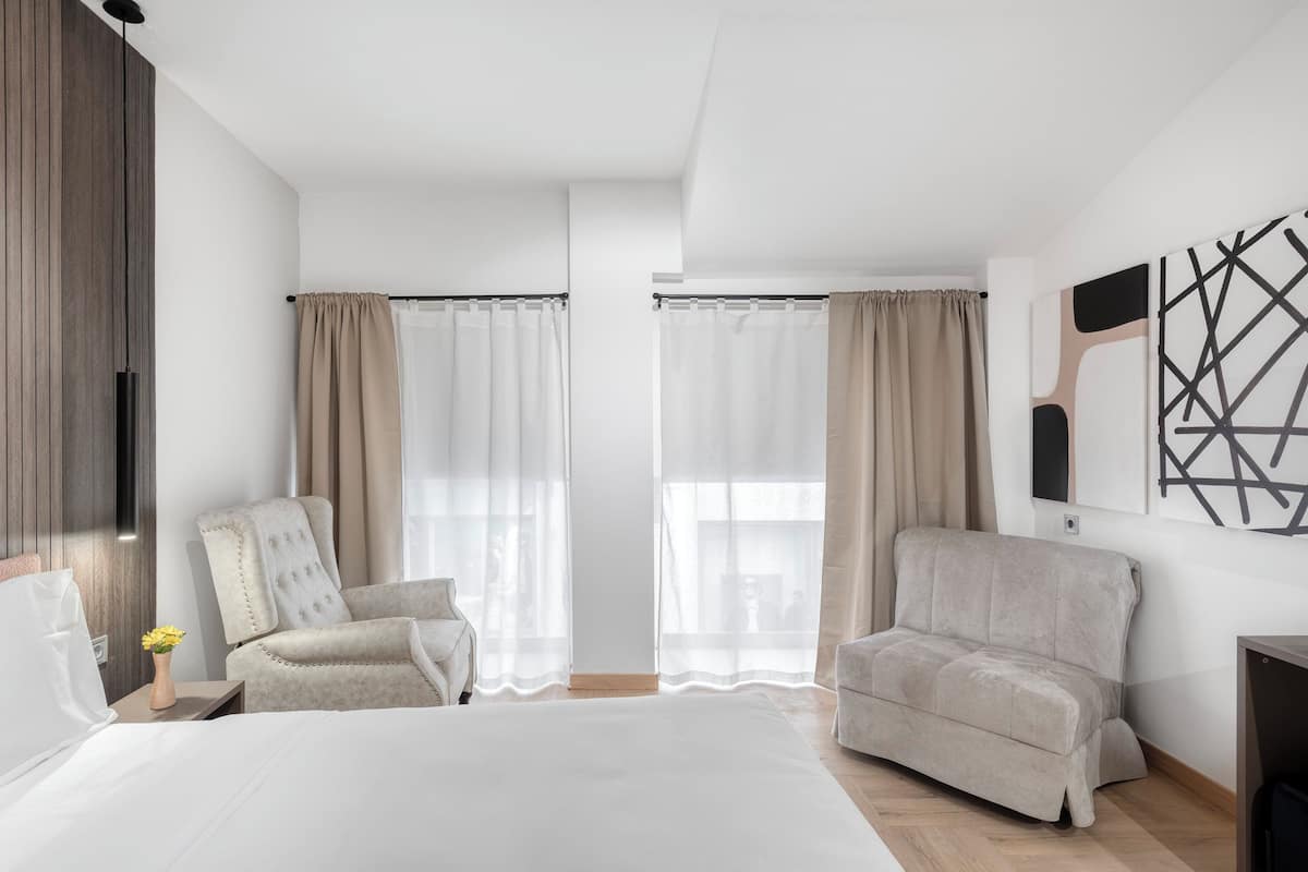 A modern hotel room with a large bed, two armchairs, a wooden panel wall, and a large window with sheer curtains.