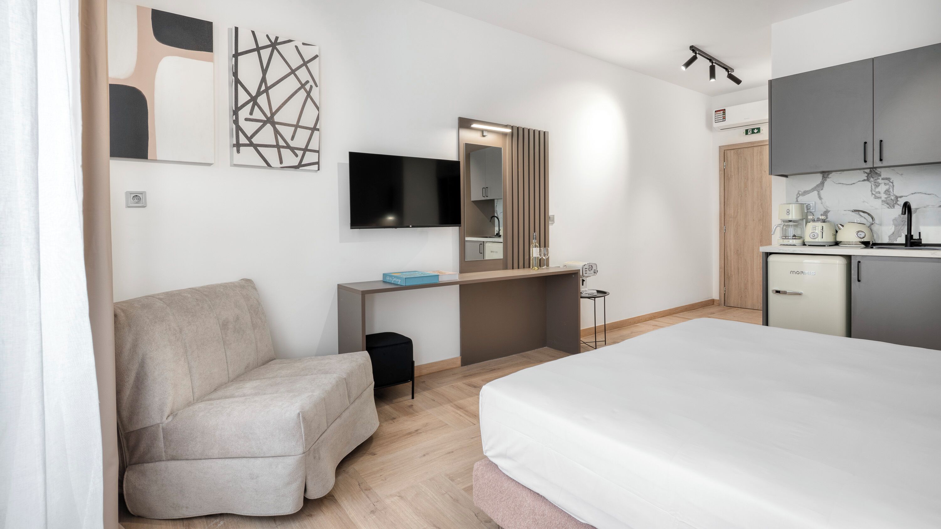 A modern hotel room with a bed, a sofa, a TV, a mirror, a kitchenette, and artwork on the walls.