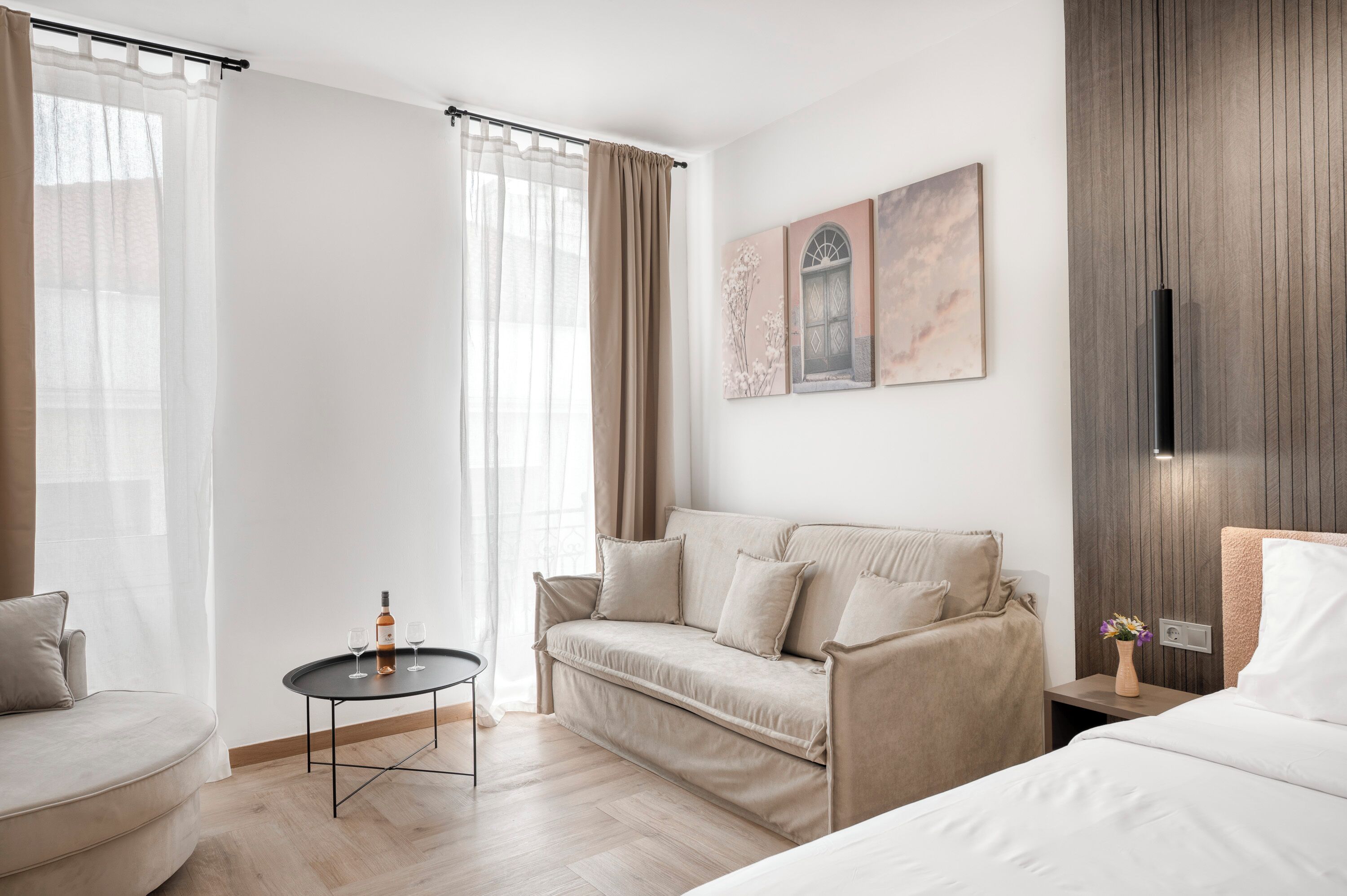 A modern hotel room with a sofa, a small round table with a bottle and glasses, a bed, a nightstand with a vase, and artwork on the wall.