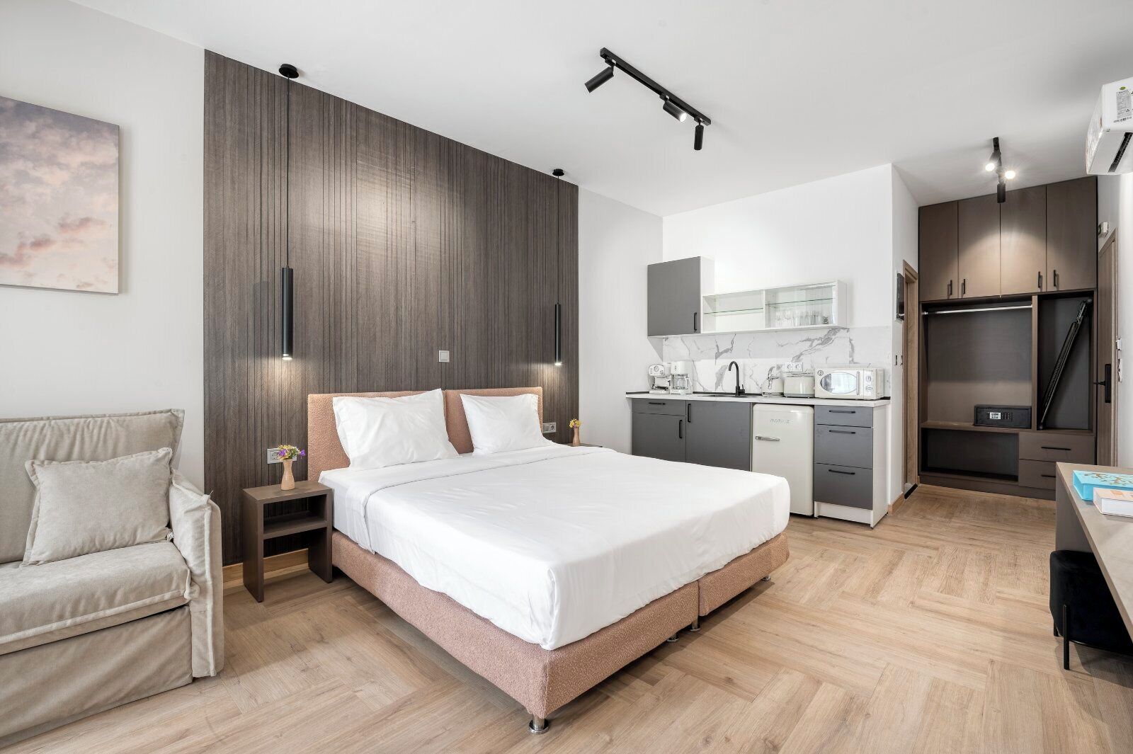 A modern bedroom with a large bed, bedside tables, a nightstand, a sofa, a built-in wardrobe, and a kitchenette.