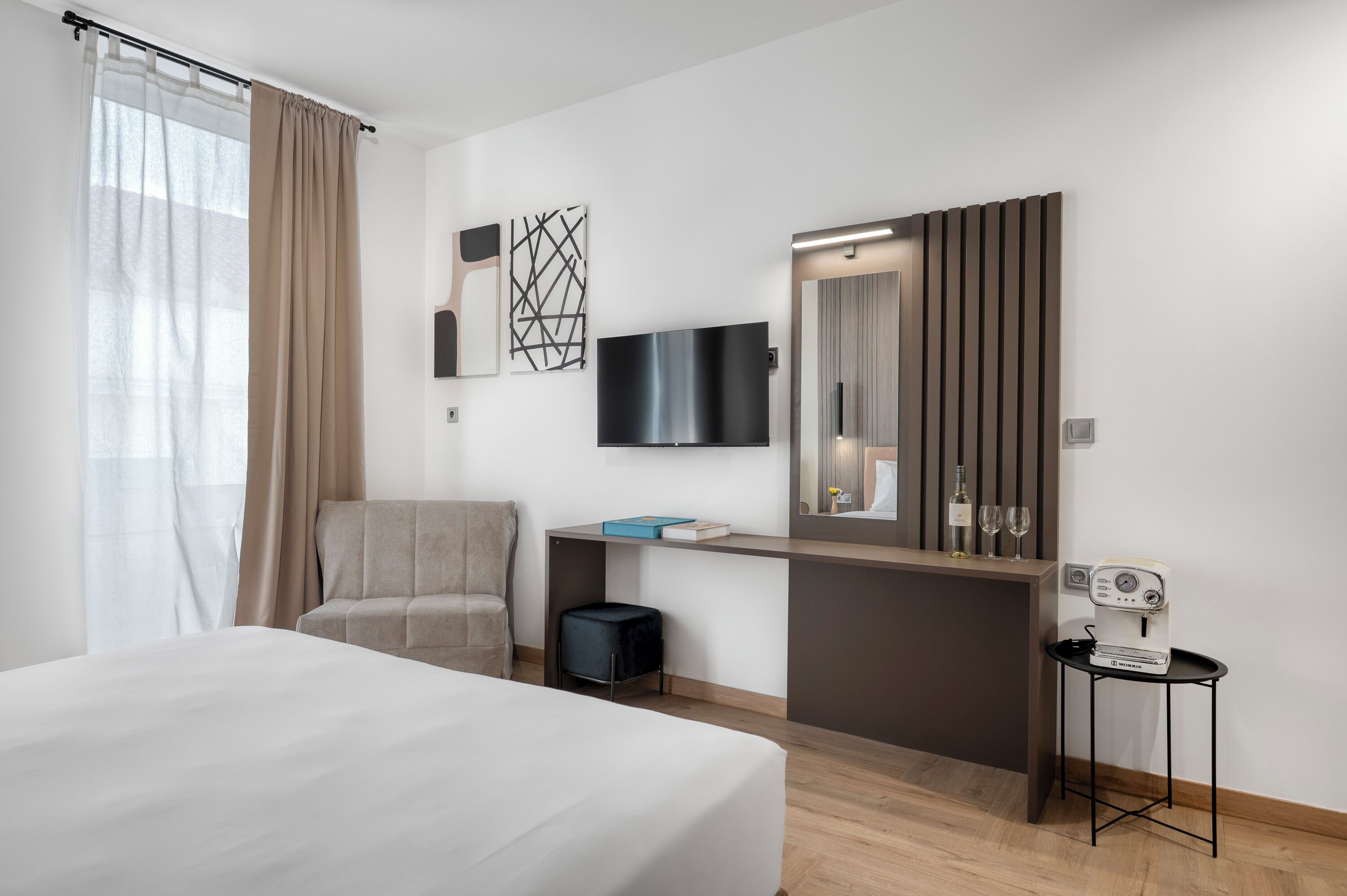 A hotel room with a bed, a chair, a desk with a flat-screen TV, a mirror, and a small table with a coffee machine.
