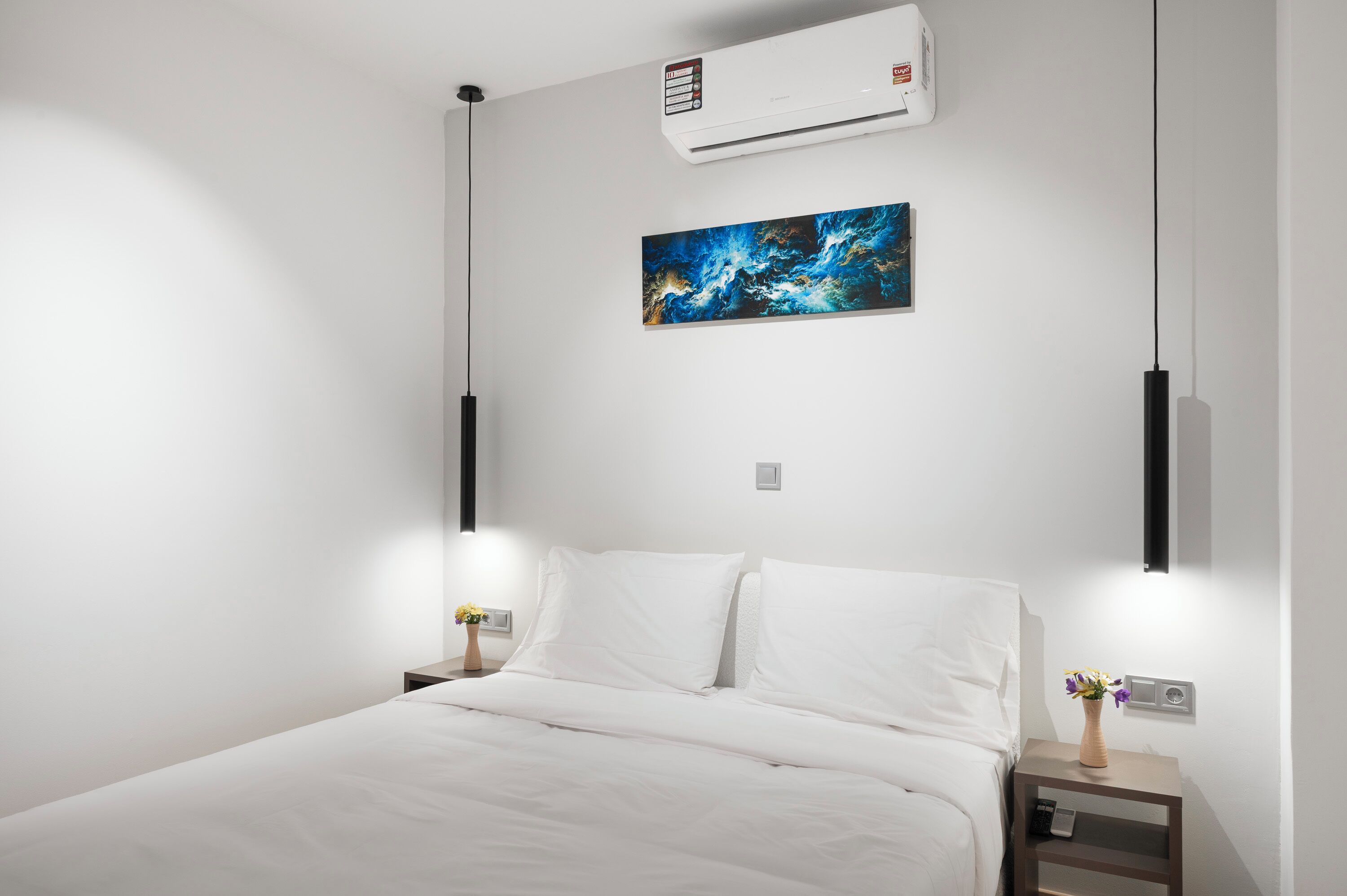 Una habitación de hotel moderna con cama, paredes blancas, aire acondicionado, un cuadro y lámparas de noche.