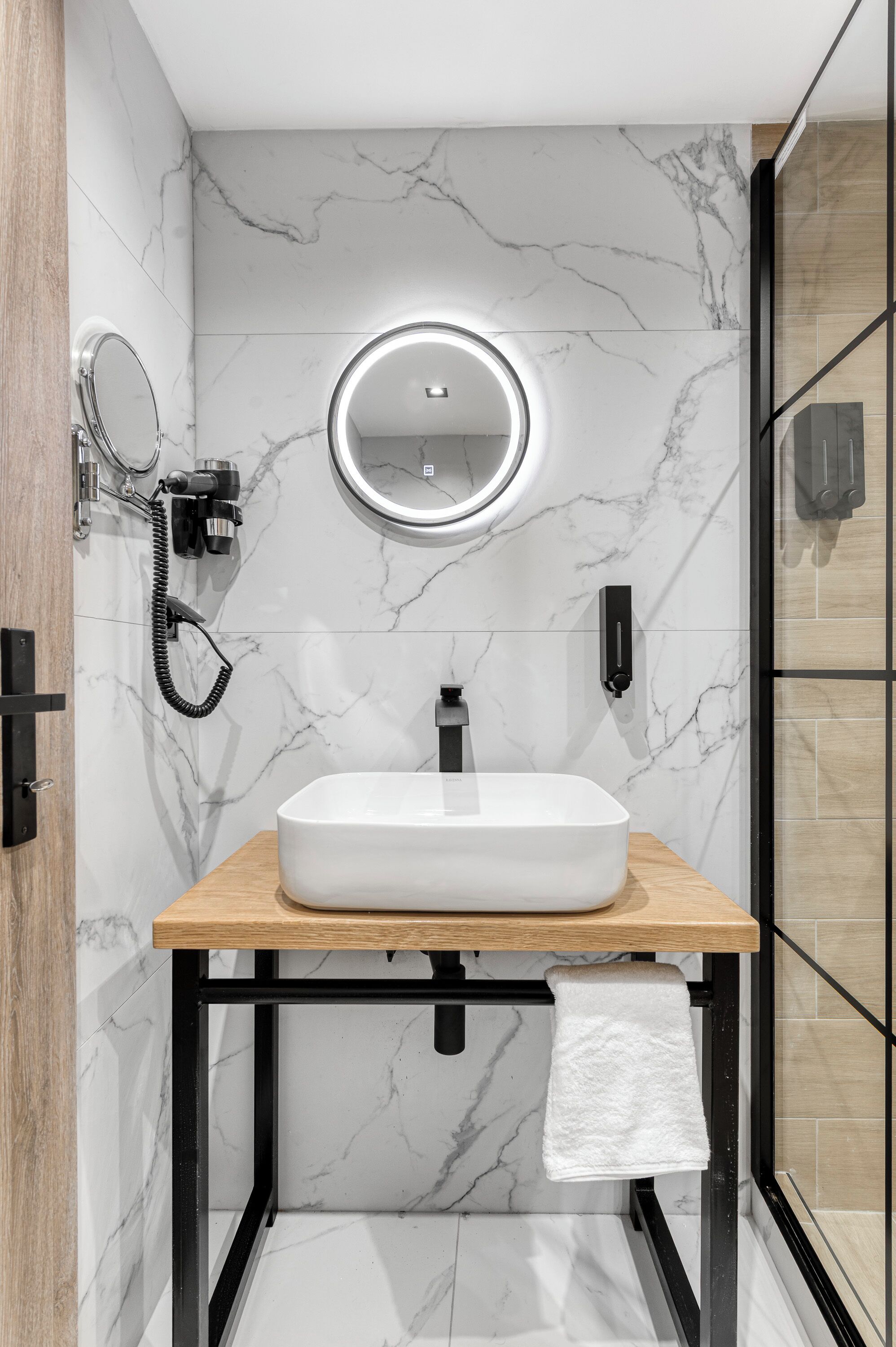 A bathroom with a marble wall, a wooden vanity, a round mirror, a handheld showerhead, and a towel hanging on a rack.