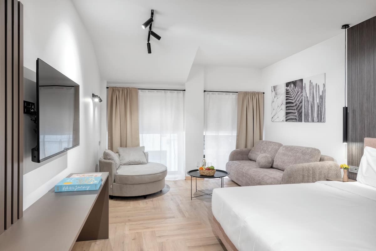 A modern hotel room with a flat-screen TV, a round armchair, a sofa, a bed, and a small table with a fruit basket.
