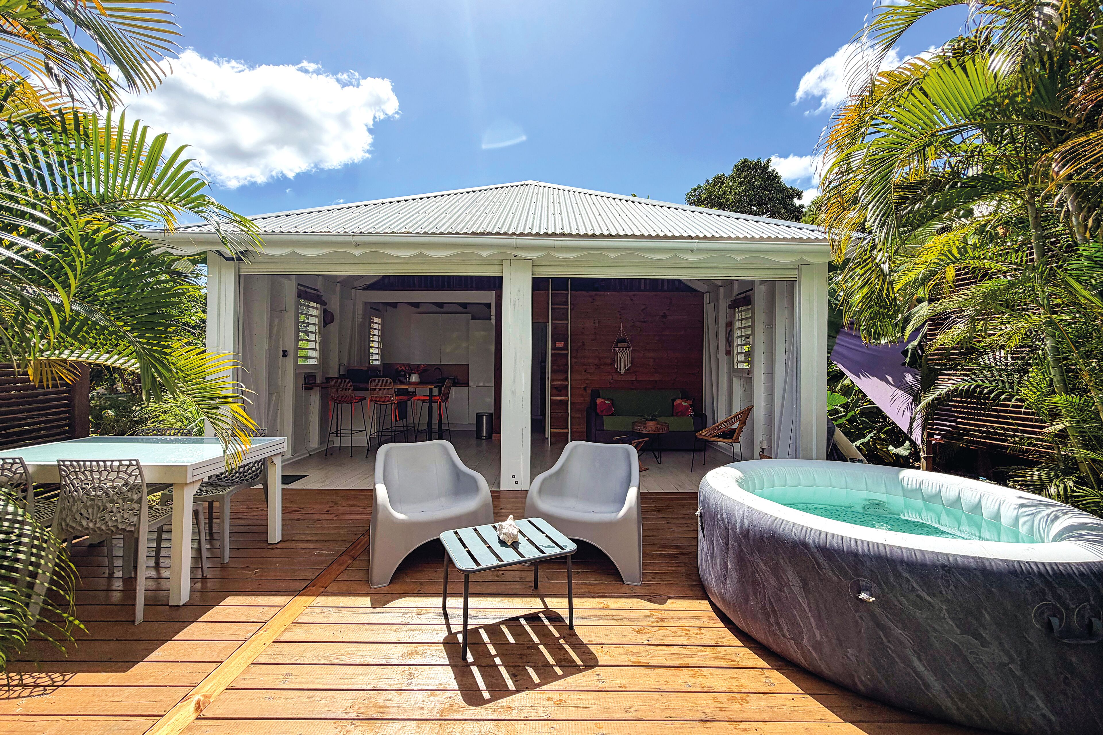 A covered outdoor area with a wooden deck, two white chairs, a small table, and a hot tub.
