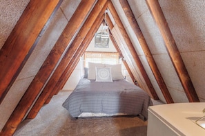 An attic bedroom with a sloped ceiling, wooden beams, a bed with a gray bedspread, and a white bedside table.
