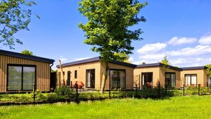 Una hilera de cabañas modernas de madera con ventanas amplias, ubicadas en una zona cubierta de césped bajo un cielo despejado.