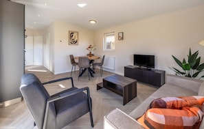 Uma sala de estar moderna com sofá, poltrona, mesa de centro, televisão, mesa de jantar com cadeiras e uma planta.