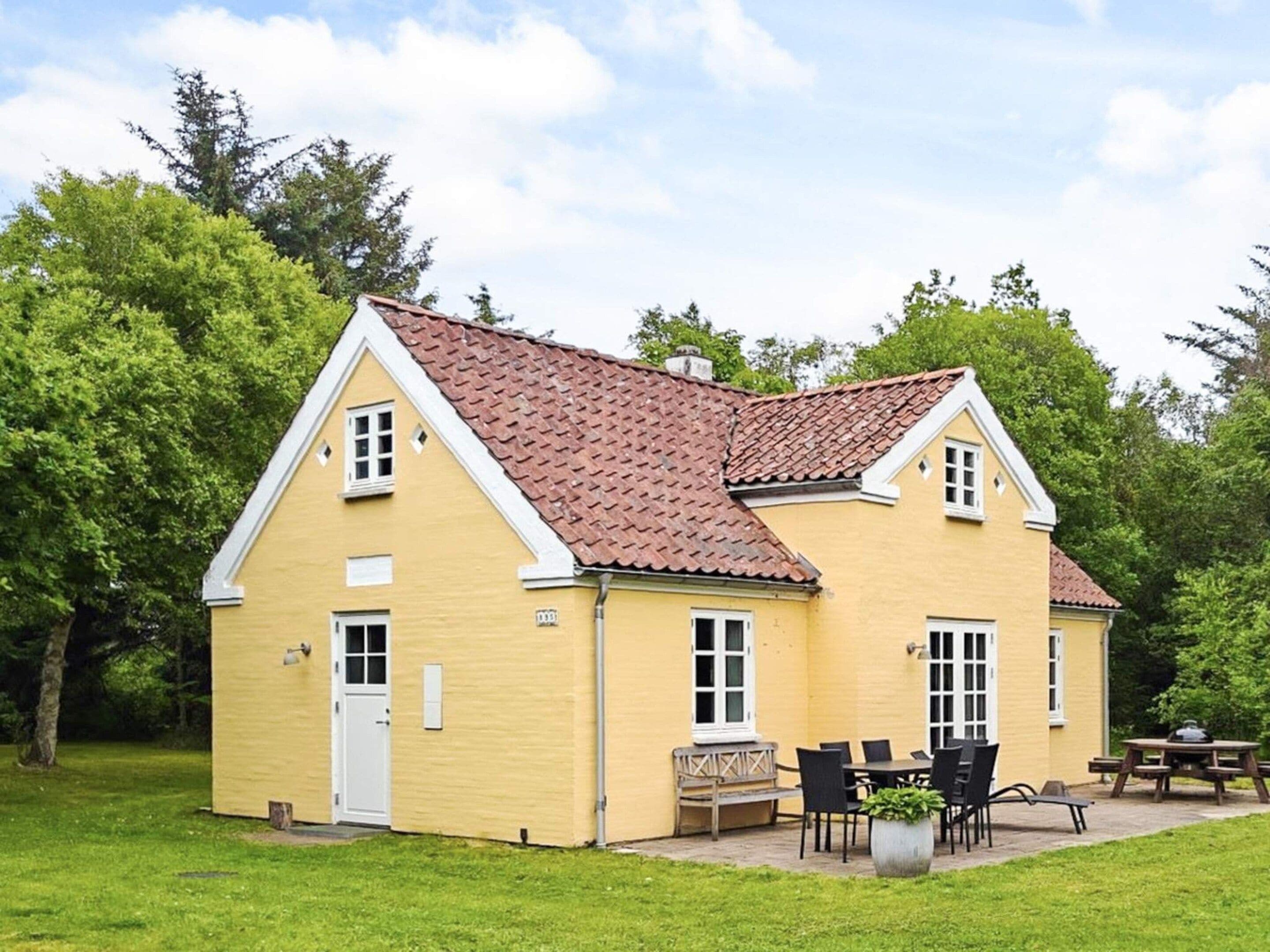 Un bâtiment jaune avec un toit recouvert de tuiles rouges, une porte d’entrée et un patio aménagé avec du mobilier d’extérieur.