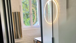 Ein Badezimmer mit Duschabtrennung aus Glas, einem weißen Waschbecken und einem Fenster mit Blick auf Grünflächen.