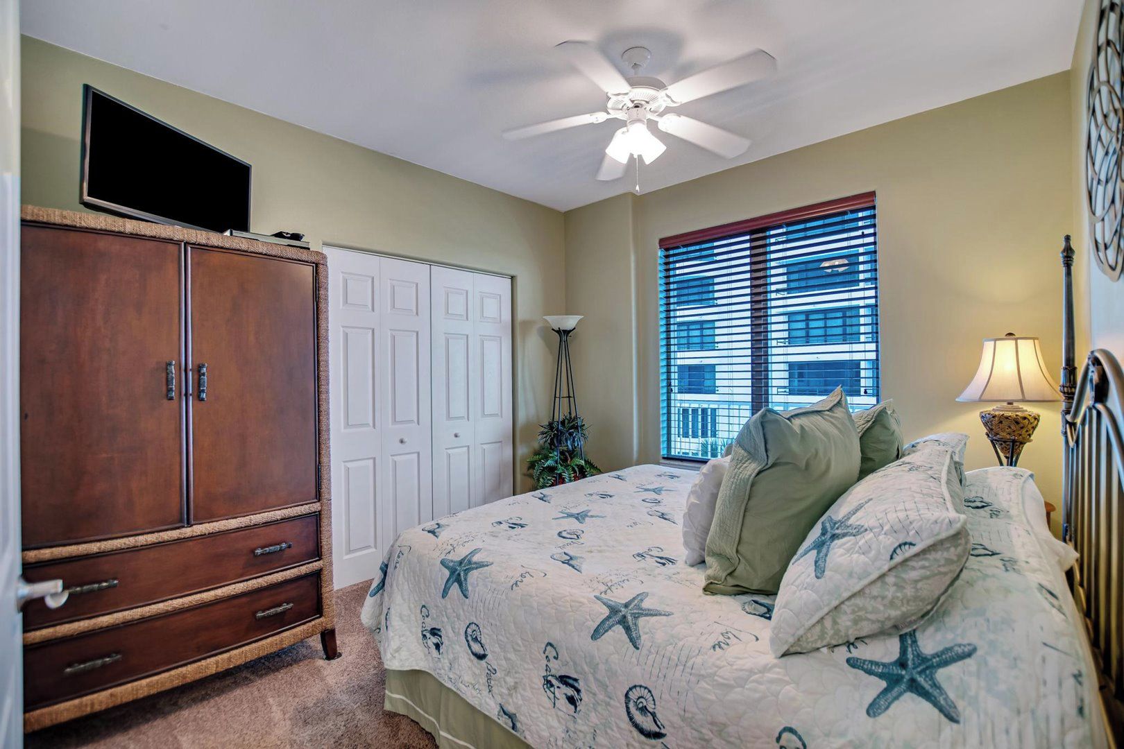 A bedroom with a bed, a wardrobe, a ceiling fan, a window with blinds, and a lamp.