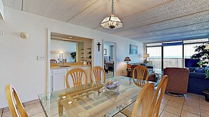 A dining area with a glass table and wooden chairs, a kitchen in the background, and a balcony with a view. - Gulf-view Resort Retreat: Suntide III - Unit 209 (South Padre Island)