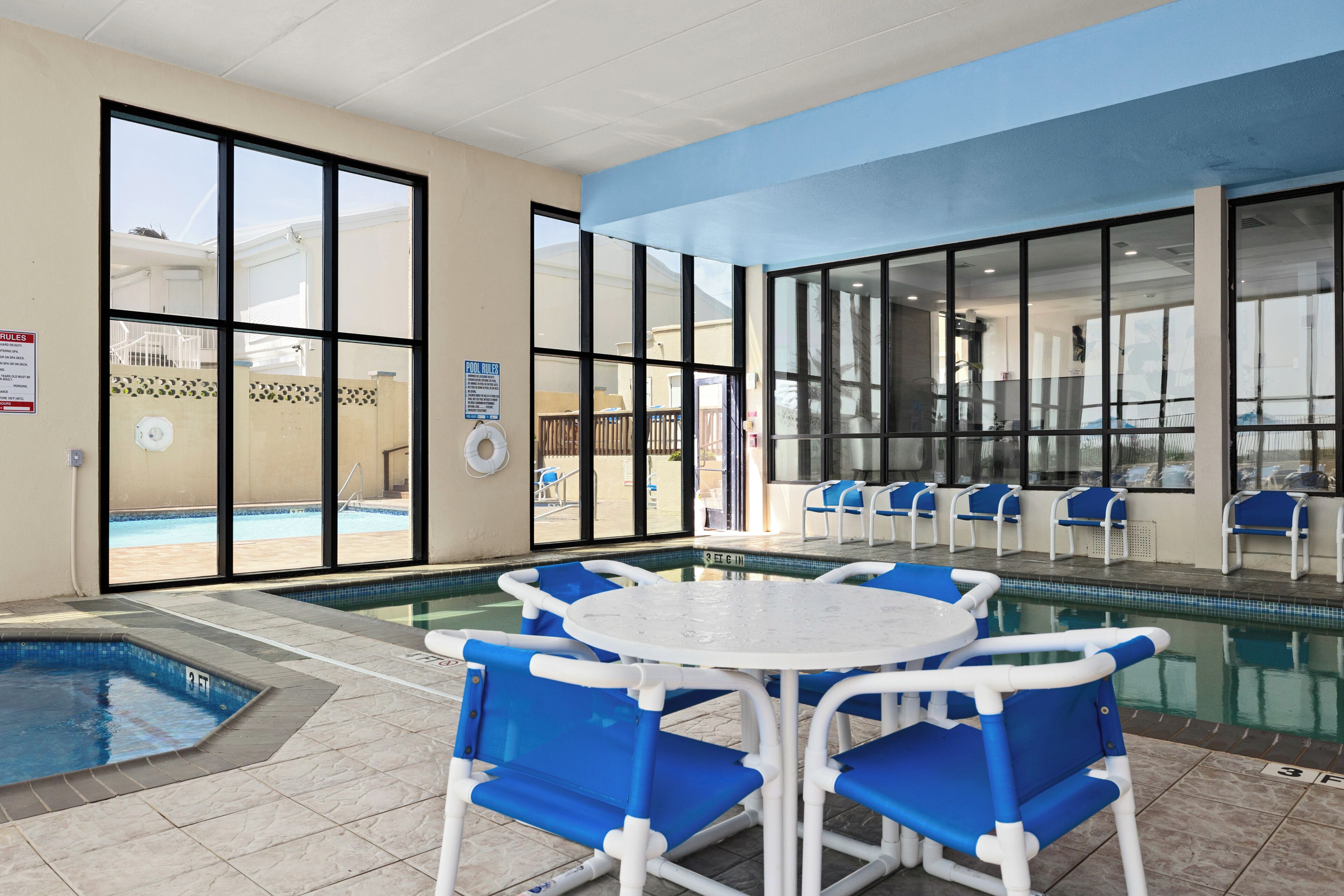 An indoor pool area with blue and white pool chairs, a small pool table, and clear glass walls.
