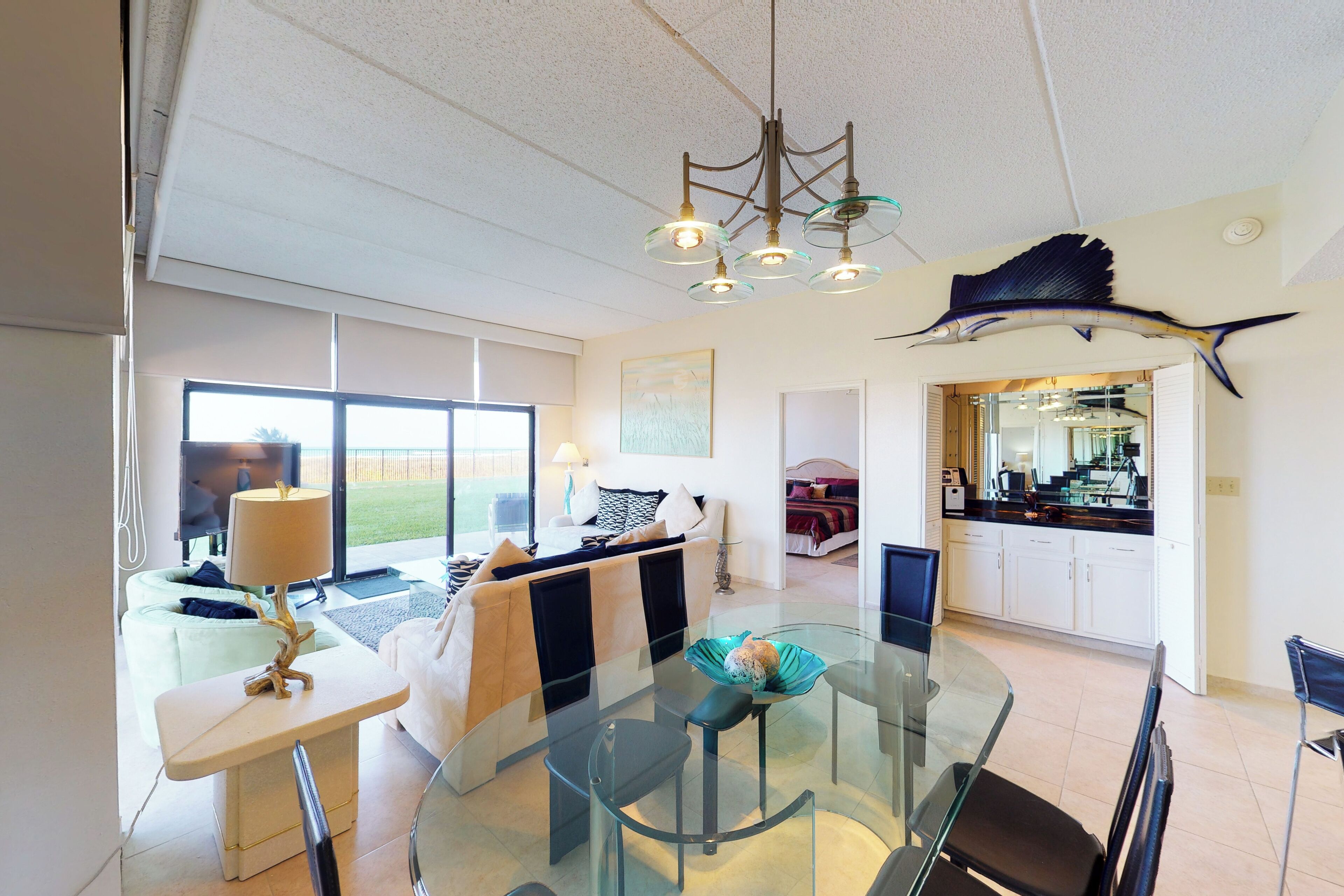 Una sala de estar moderna con una mesa de comedor de vidrio, un sofá y una cocina visible a través de un umbral abierto.