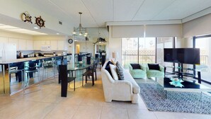 Een moderne keuken met glazen kastjes, een eethoek met stoelen, een bank, een televisie en uitzicht op een balkon.