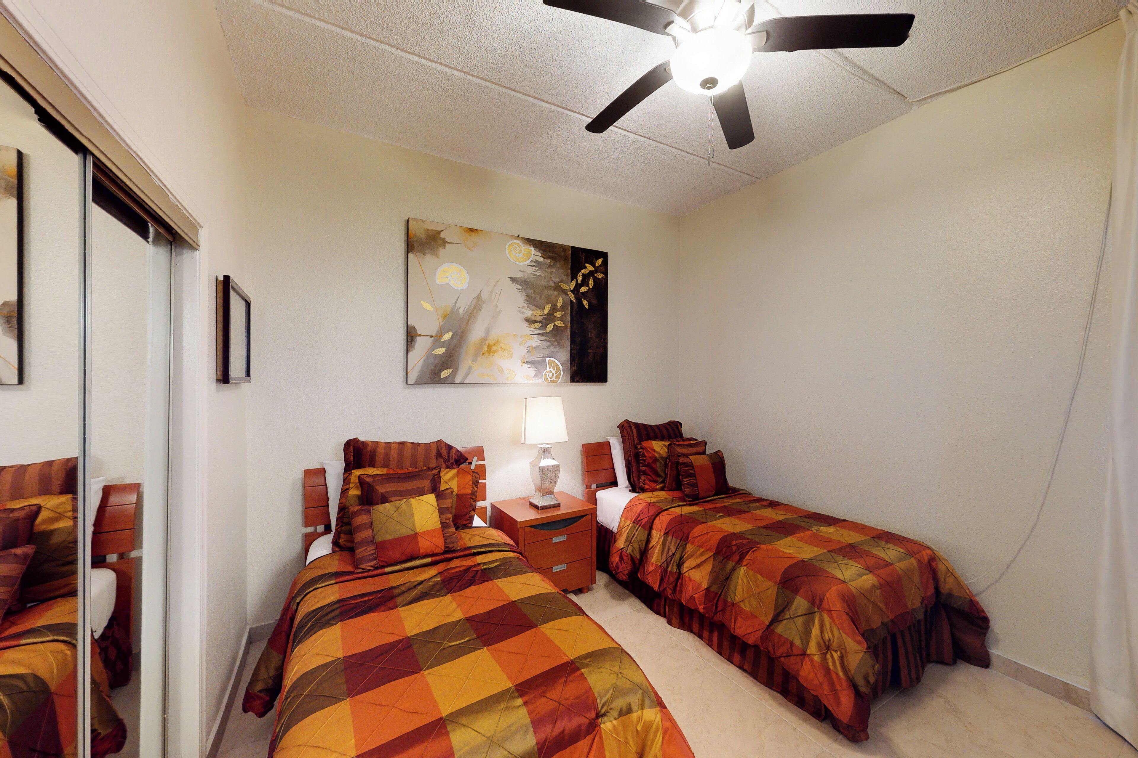 A bedroom with two beds, a ceiling fan, a nightstand, a lamp, and a painting on the wall.