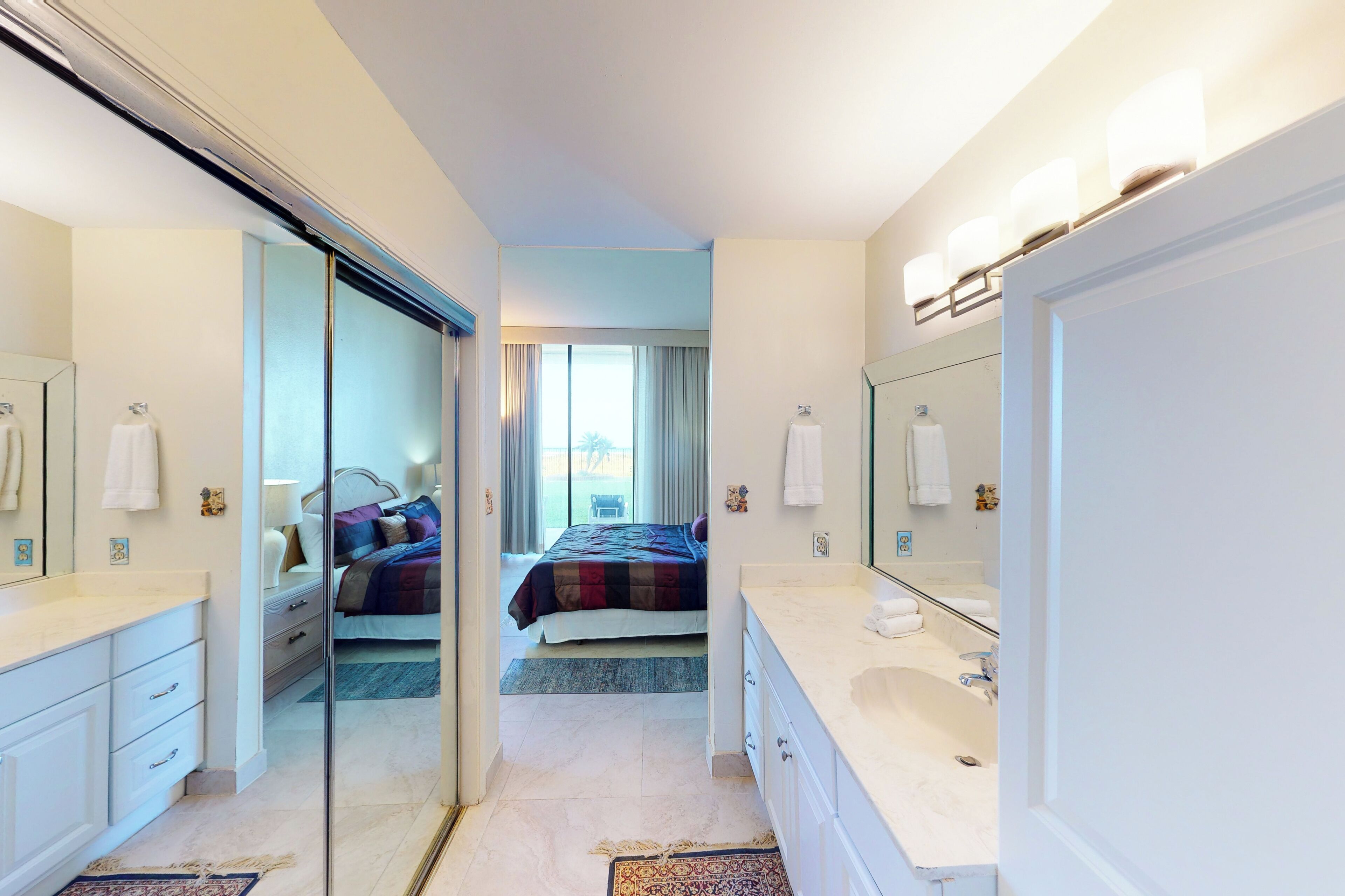 A hotel room with a large mirror, a double bed, a bathroom with a sink and mirror, and a view of the outside.