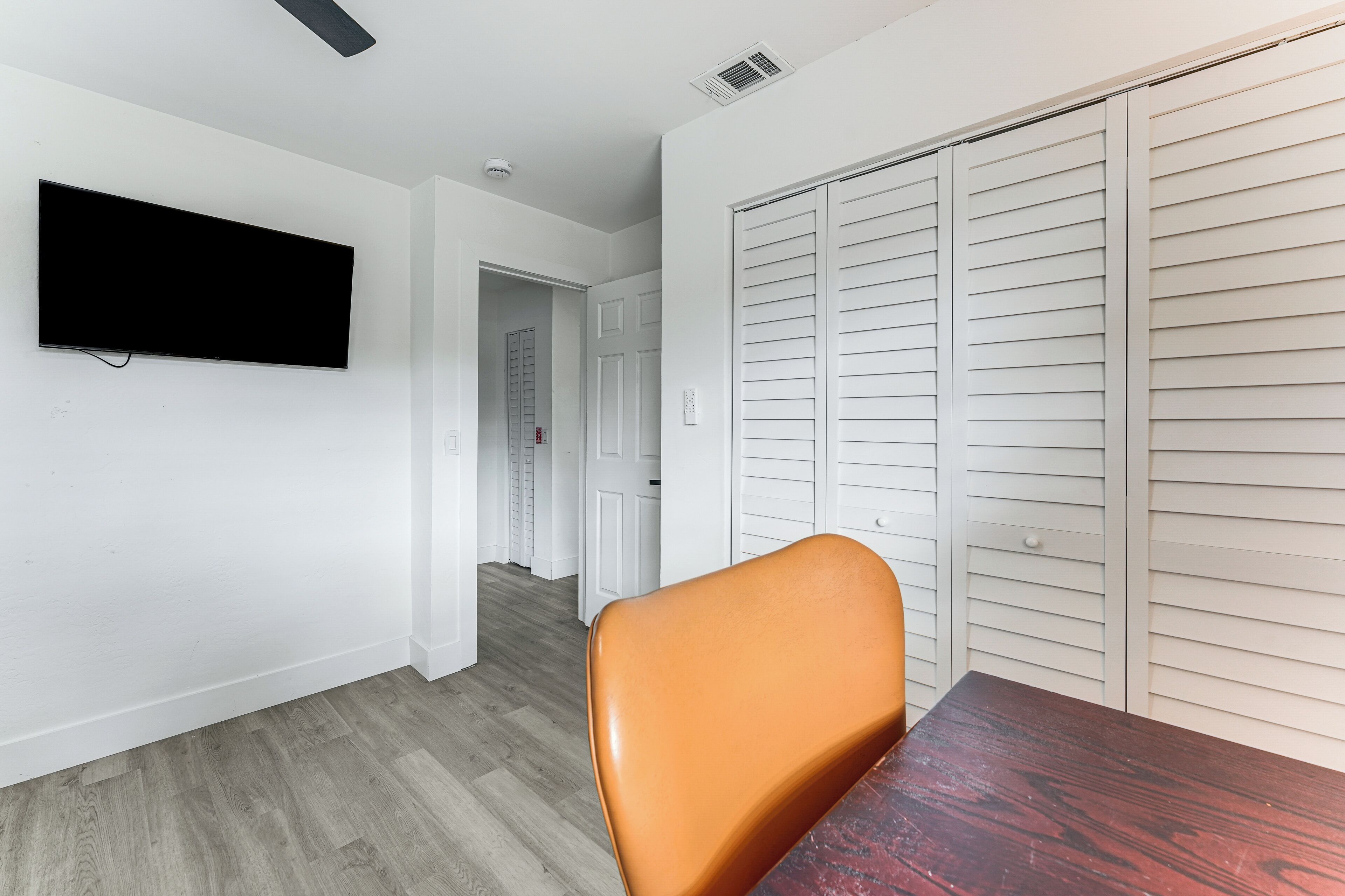 A room with a flat-screen TV mounted on the wall, a wooden dining table, an orange chair, and a white closet with horizontal louvered doors.