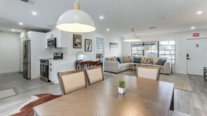 A modern kitchen with stainless steel appliances, a dining area with a wooden table and chairs, and a living room with a sofa and a television.