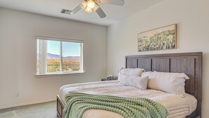 A bedroom with a large bed, a ceiling fan, a window with a view of mountains, and a painting on the wall.