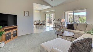 A spacious living room with a flat-screen TV, a ceiling fan, a dining area with chairs and a table, and large windows offering a view of the outdoors.