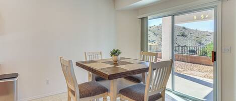 Un coin repas avec une table et des chaises en bois, une plante en pot et une vue sur un espace extérieur aménagé, visible à travers une porte-fenêtre coulissante.