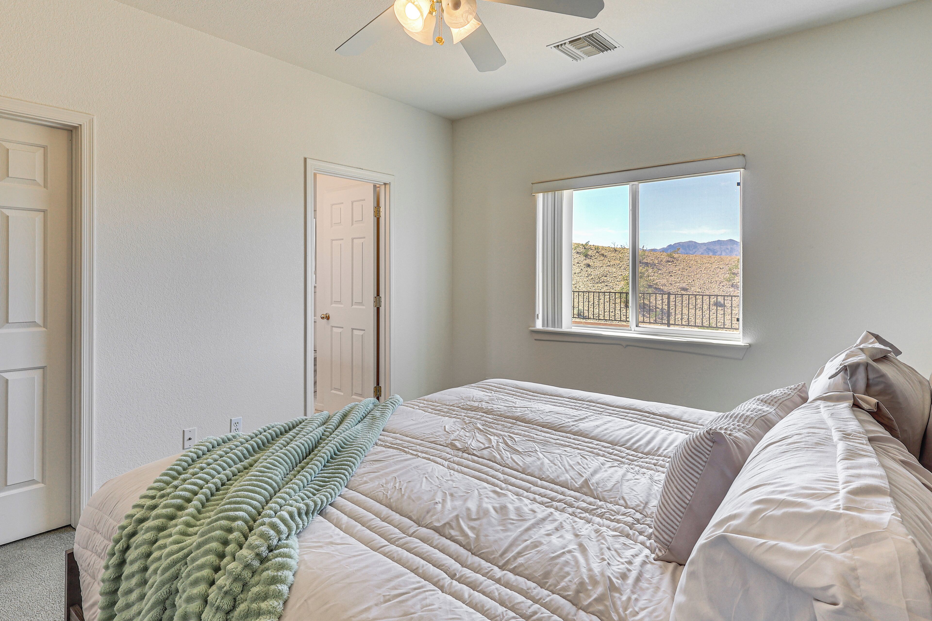 A bedroom with a bed, a window with a view of a desert landscape, a ceiling fan, and a door.