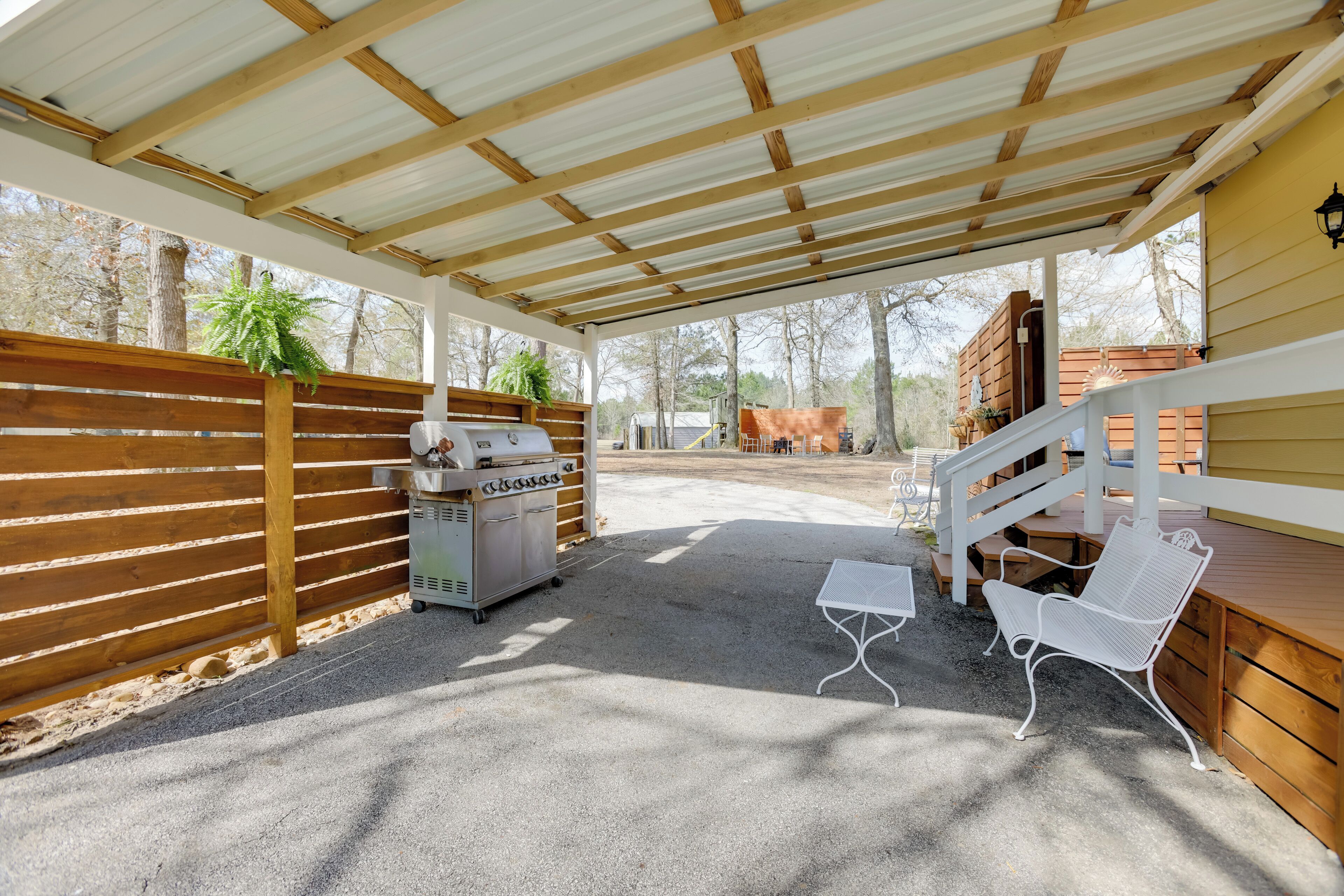 Un patio techado al aire libre con una parrilla, sillas de metal blanco y una cerca de madera.
