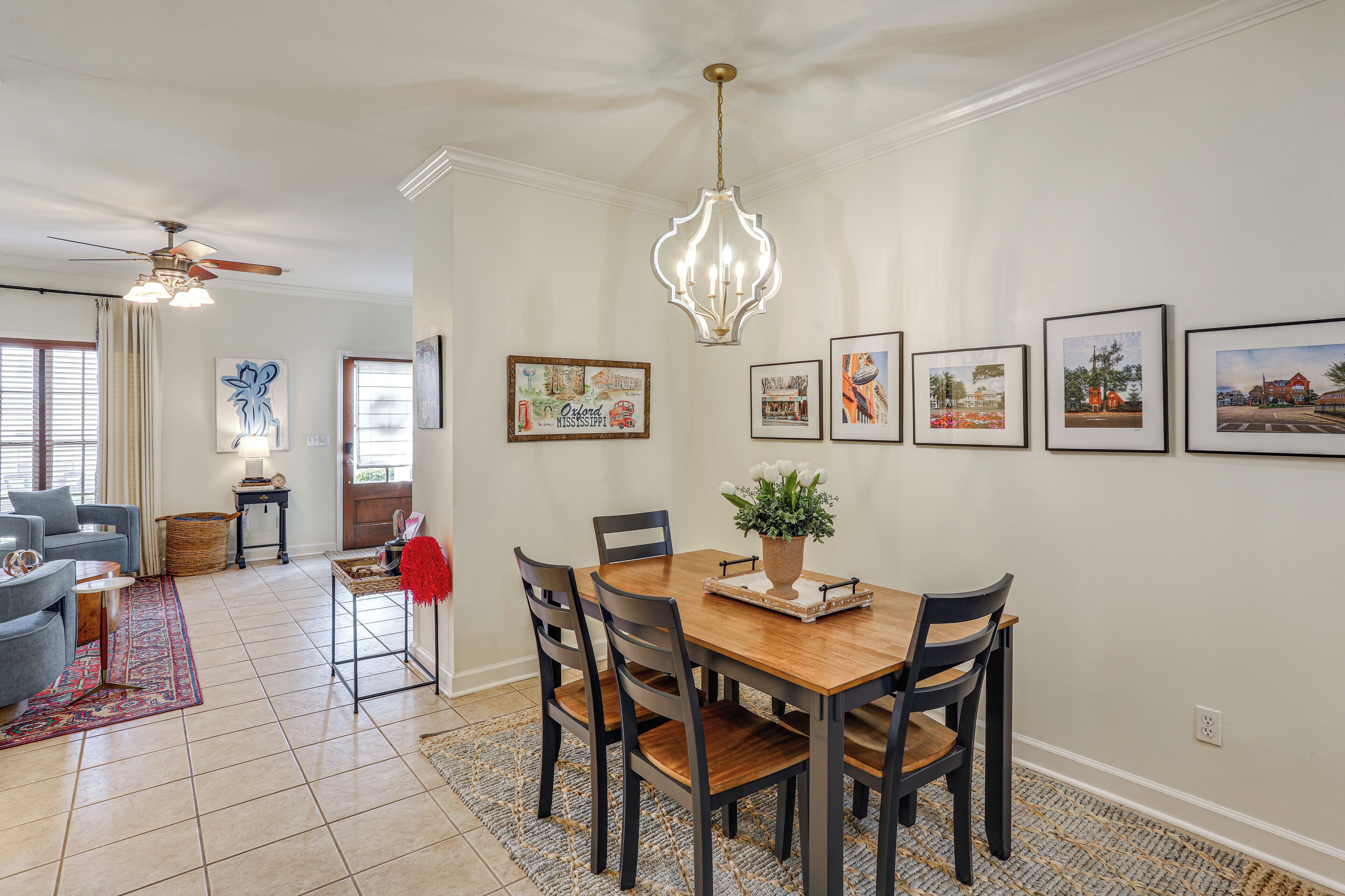 A dining area with a wooden table, chairs, and a chandelier. There are framed pictures on the wall and a ceiling fan above.