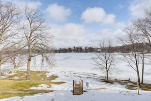 Een besneeuwd landschap met een houten bruggetje dat naar een bevroren meer leidt, omringd door kale bomen.