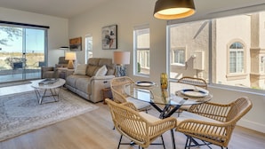 A modern living room with a glass coffee table, wicker chairs, a sofa, and a large window offering a view of a building. - Private Golf Cart! 55-plus Green Valley Getaway (Green Valley)