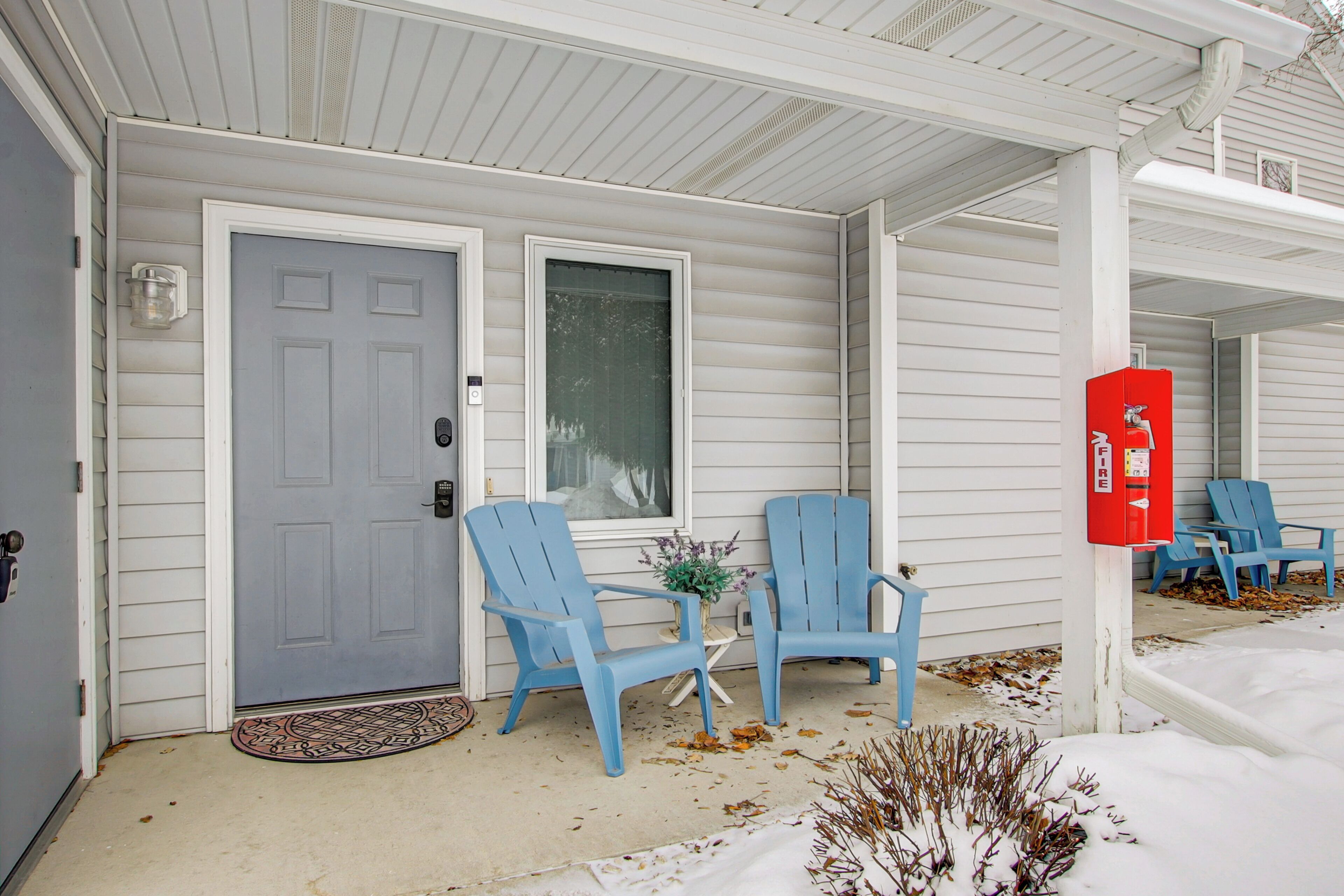 A front porch with a gray door, two blue Adirondack chairs, a small table with a vase, and a red fire extinguisher.