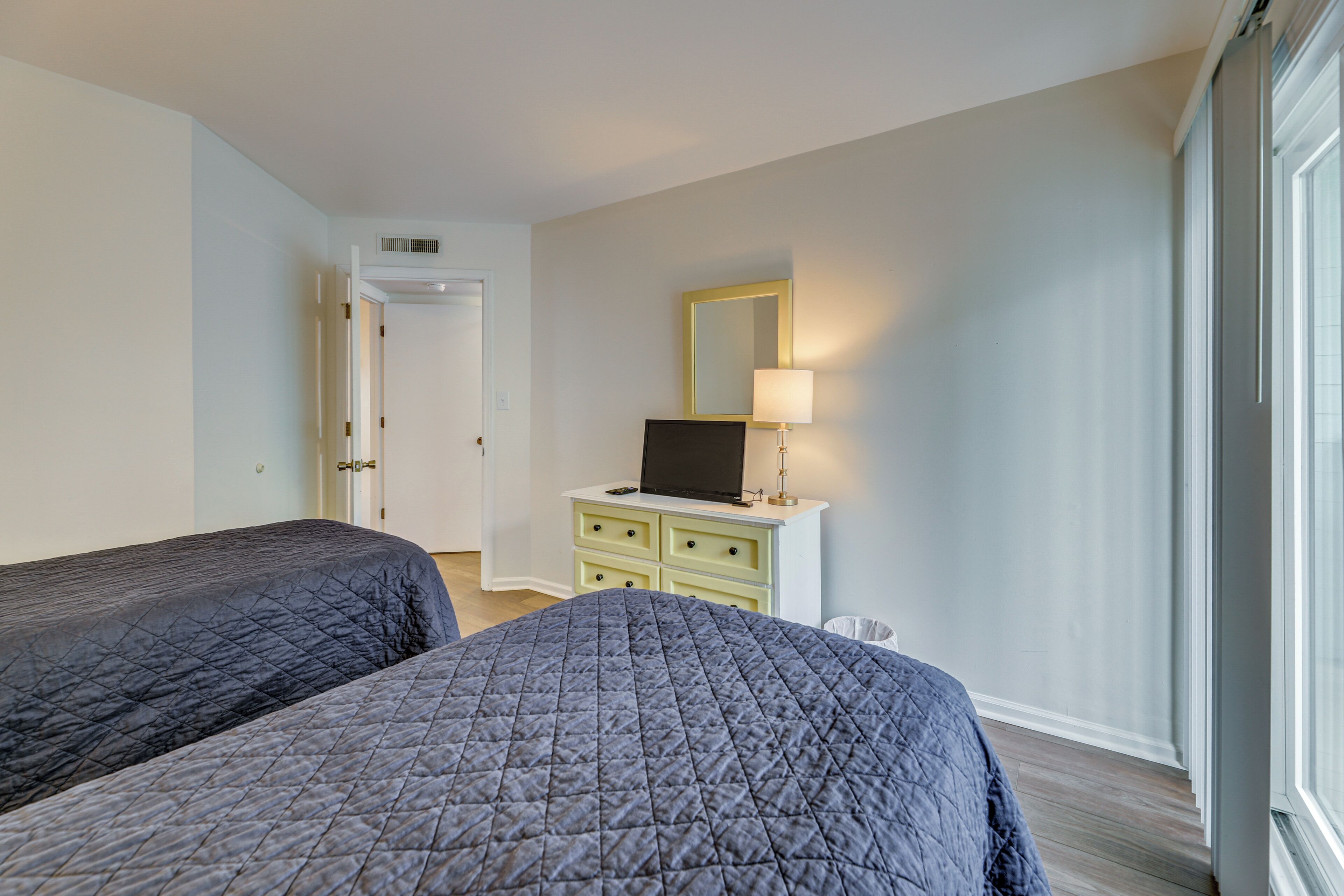 A hotel room with two beds, a desk with a lamp, a mirror, and a window.