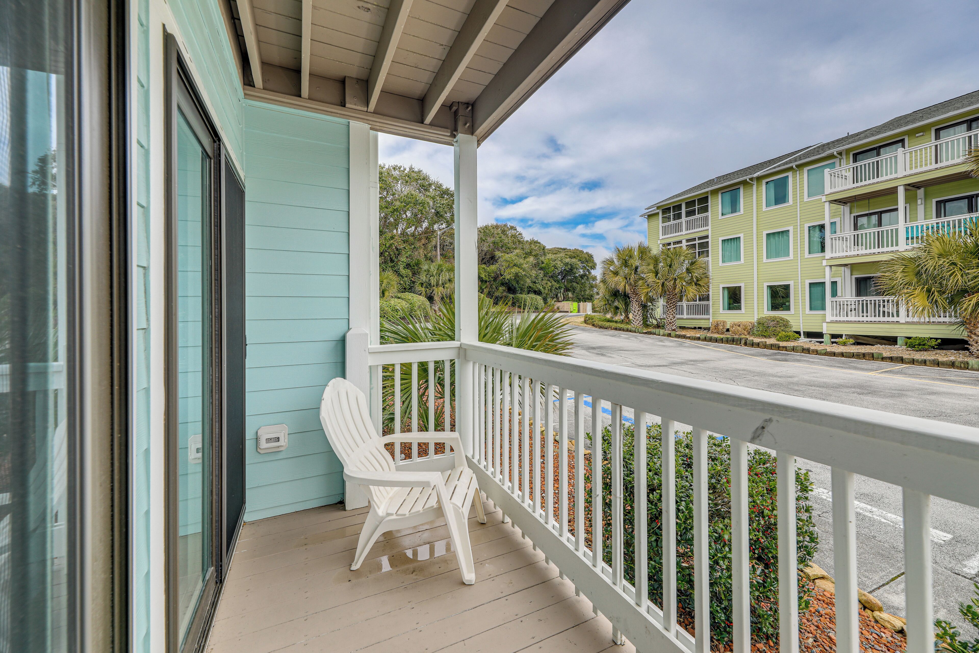 A porch with a white chair, a railing, and a view of a building with multiple balconies and greenery.