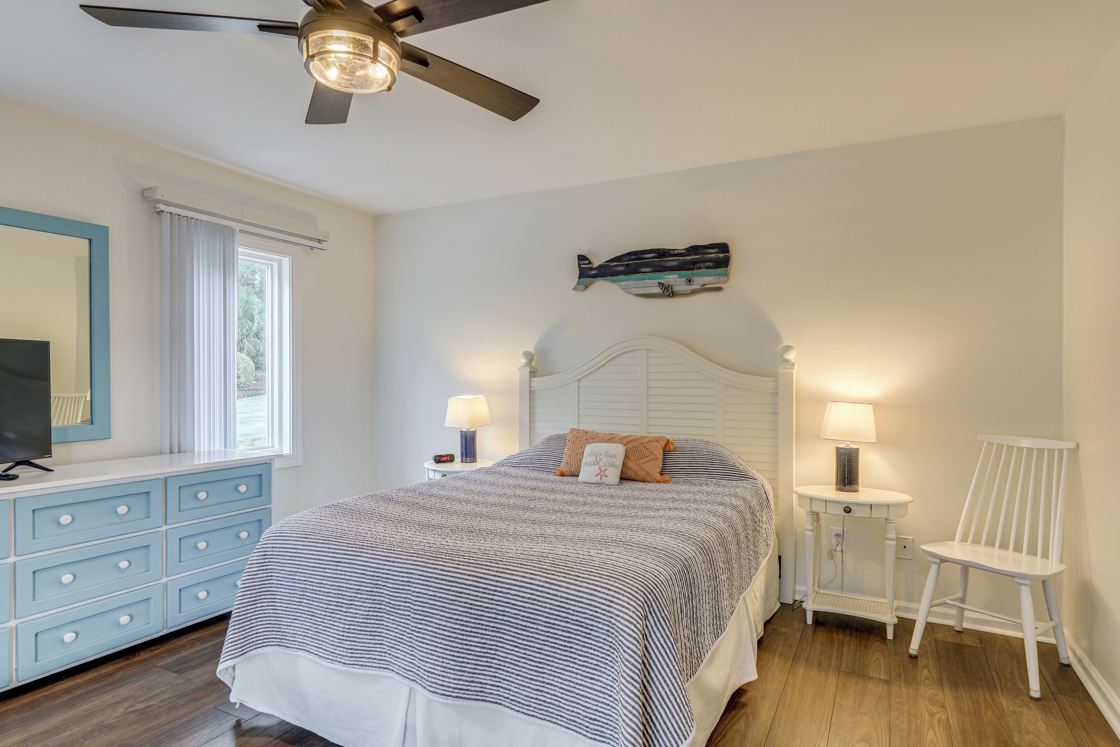 A bedroom with a bed, a dresser, a chair, a nightstand, a lamp, a ceiling fan, and a wall art piece.