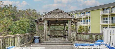 A wooden deck with blue and white lounge chairs, a gazebo, and a building with balconies in the background.